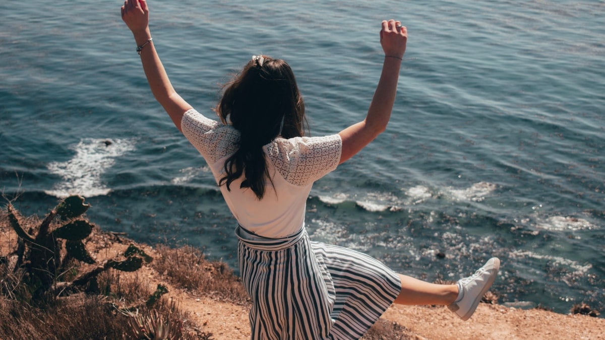 woman in white and black stripe dress standing on brown rock near body of water during